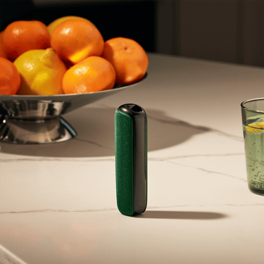 جهاز Ploom مع غطاء خلفي باللون الأخضر الأمازوني، على طاولة بالقرب من سلة فواكه.