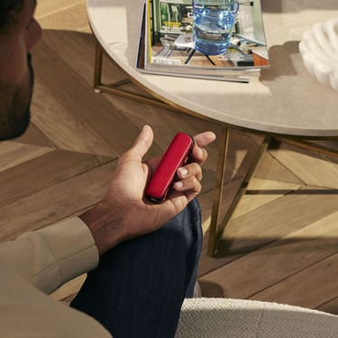Man holding a Ploom device with a red back cover while sitting on a chair near a coffee table with magazines and a glass of water.